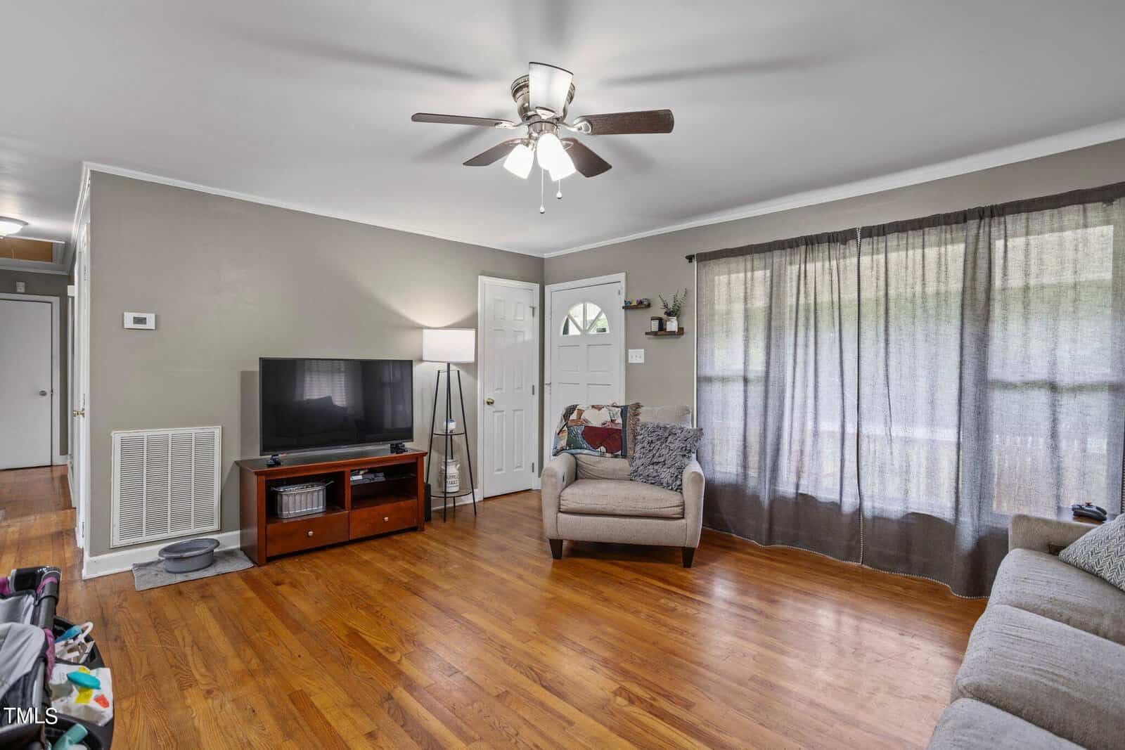 Living room with wooden floor and light green walls. A ceiling fan is above. A TV on a stand with shelves holds a plant. Armchair with patterned pillow near a window with sheer curtains. Door on the left, and a hallway is visible.