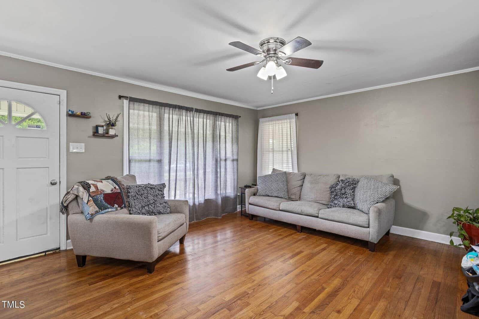 A living room with hardwood floors, featuring a ceiling fan with a light fixture. There are two gray couches, one against a window with sheer curtains. A small plant is in the corner near the couch, and a throw blanket is draped on the right couch.