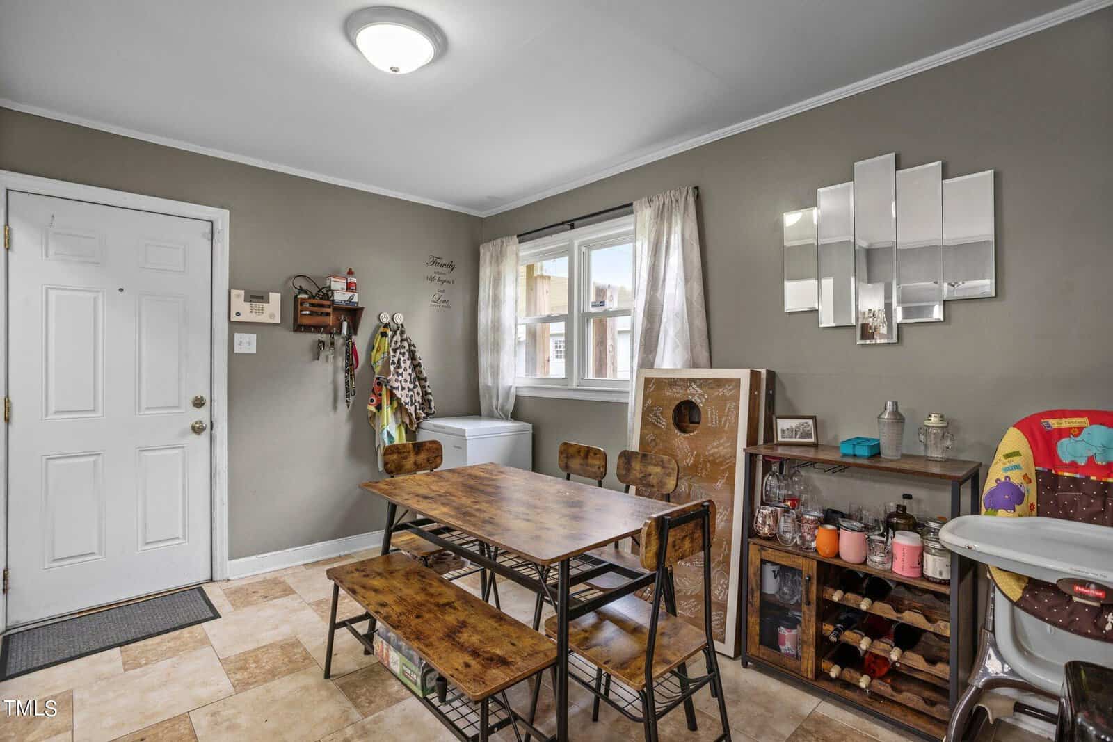 A small dining area with a wooden table and four chairs. A high chair is on the right, and a series of asymmetrical mirrors hangs on the wall. Shelves hold various jars and items. A white door and a window with curtains are on the left.