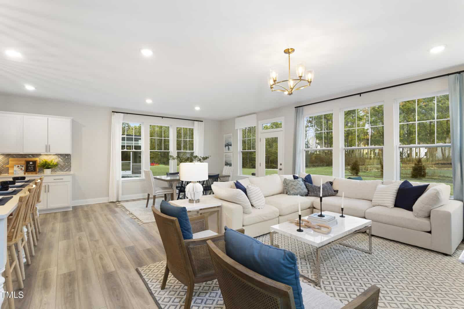 A spacious, modern living area with a beige sectional sofa, blue accent pillows, two armchairs, large windows with views of trees, and an open kitchen with white cabinets and a breakfast bar.