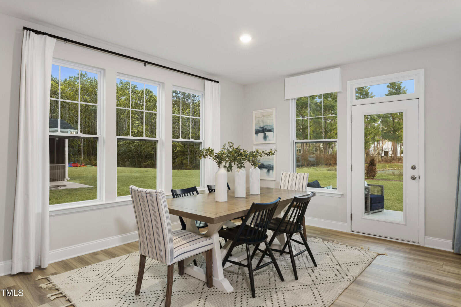 A bright dining area with large windows, a wooden table, mixed black and striped chairs, white vases with greenery, a patterned rug, and a glass door opening to a green backyard.
