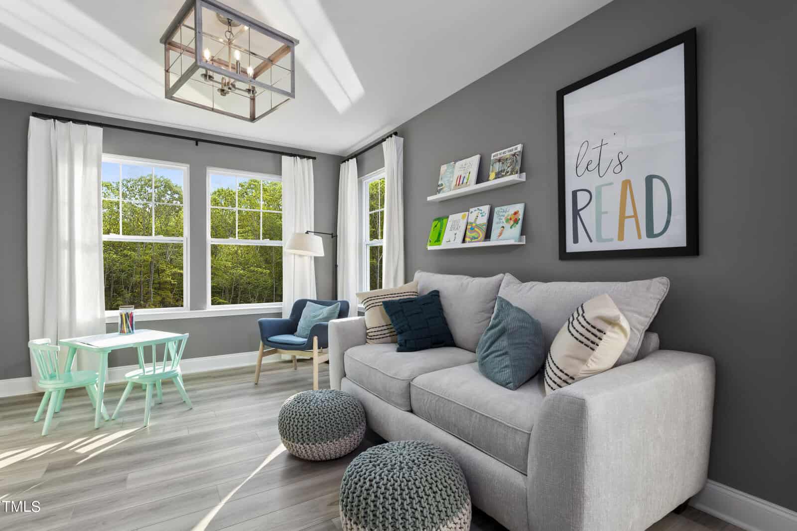 Bright reading nook with a gray sofa, teal and gray pillows, two poufs, a light wood floor, childrenโs table and chairs, books on floating shelves, and large windows showing green trees outside.
