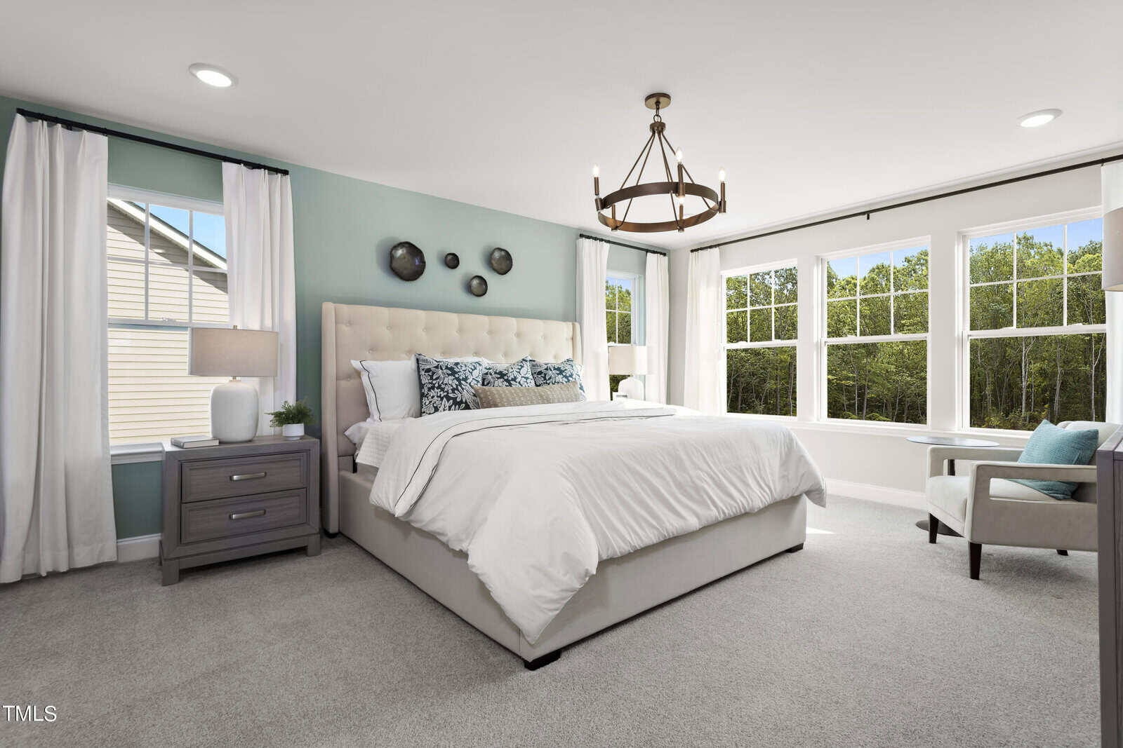 Bright bedroom with large windows, a beige upholstered bed with white bedding, a nightstand with a lamp, wall art above the bed, and a cozy armchair by the window.