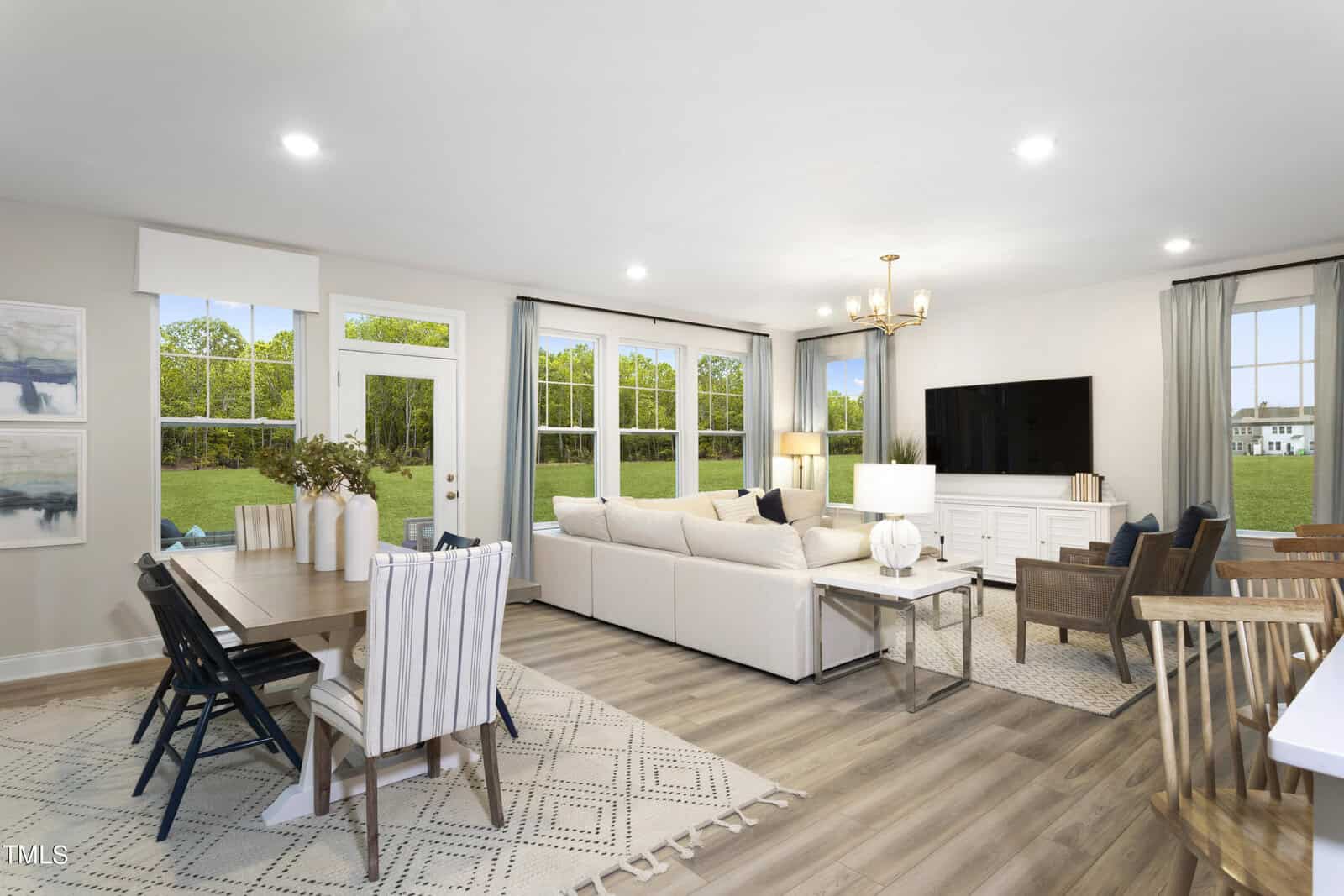 Bright, modern open-concept living and dining room with light wood floors, beige sectional sofa, TV, dining table with chairs, large windows, and a view of a green lawn and trees outside.
