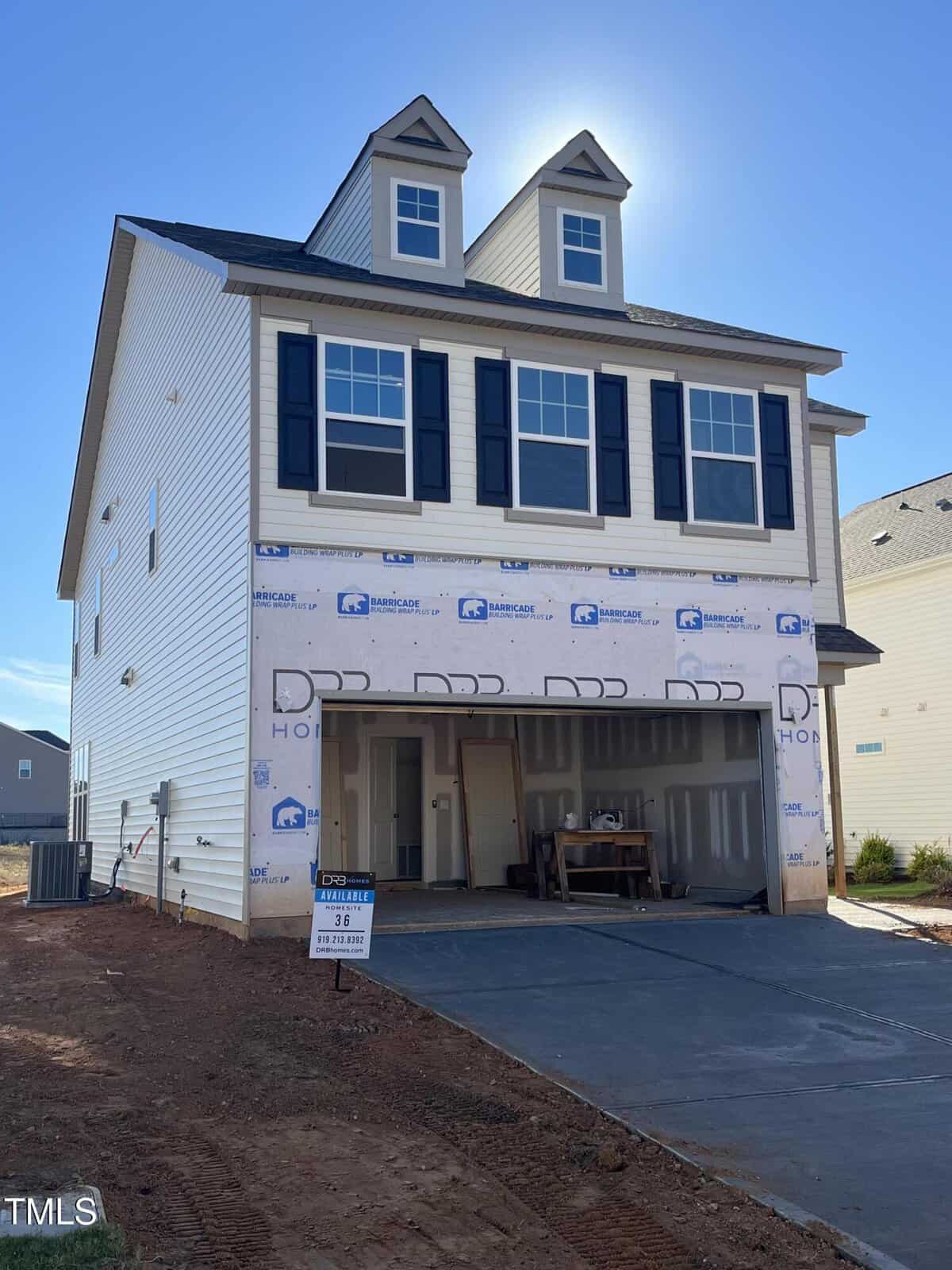 A newly constructed three-story house with white siding, black shutters, and two dormer windows. The garage is open, revealing construction materials inside. The driveway and yard are unfinished, with some exposed red soil.
