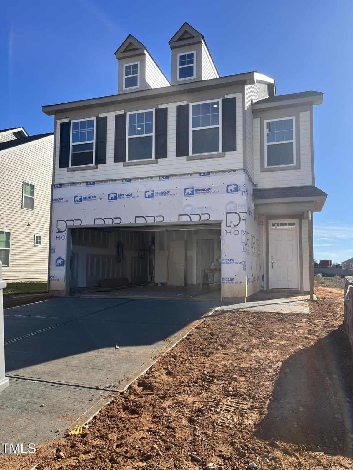 A two-story house under construction with white siding and black shutters, featuring a double garage and dormer windows. The driveway is paved, while the yard is bare dirt. Blue sky in the background.