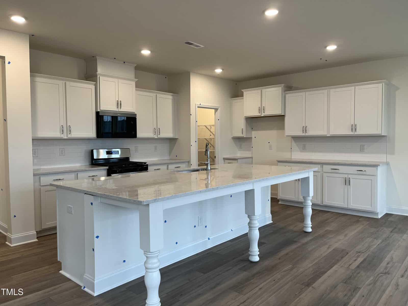 A spacious modern kitchen with white cabinets, stainless steel appliances, a large center island with a marble countertop, and wood-look flooring under bright recessed lighting.