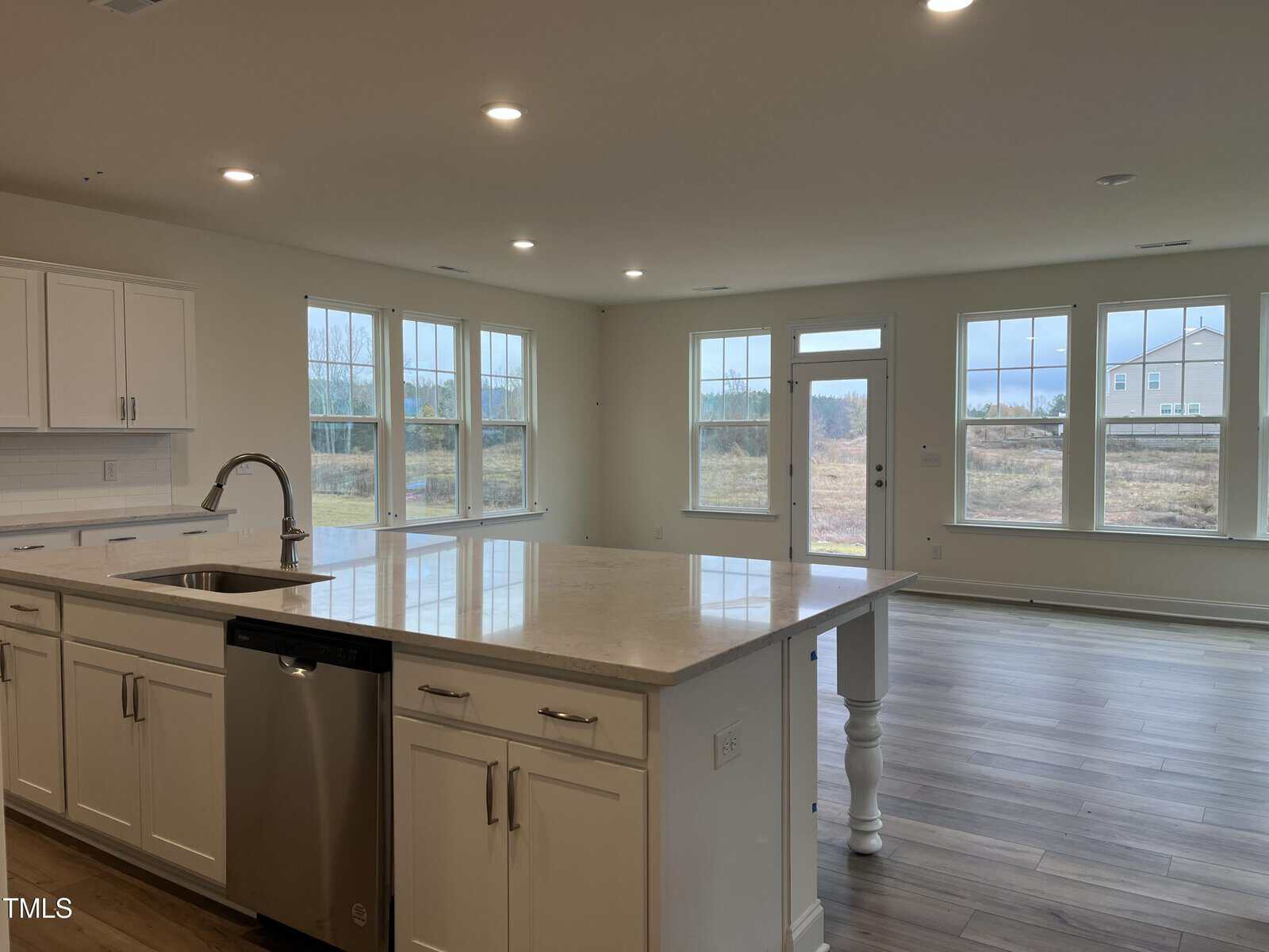 Modern open-concept kitchen and living area with white cabinets, a large island, stainless steel appliances, wood floors, and large windows overlooking a grassy outdoor area.