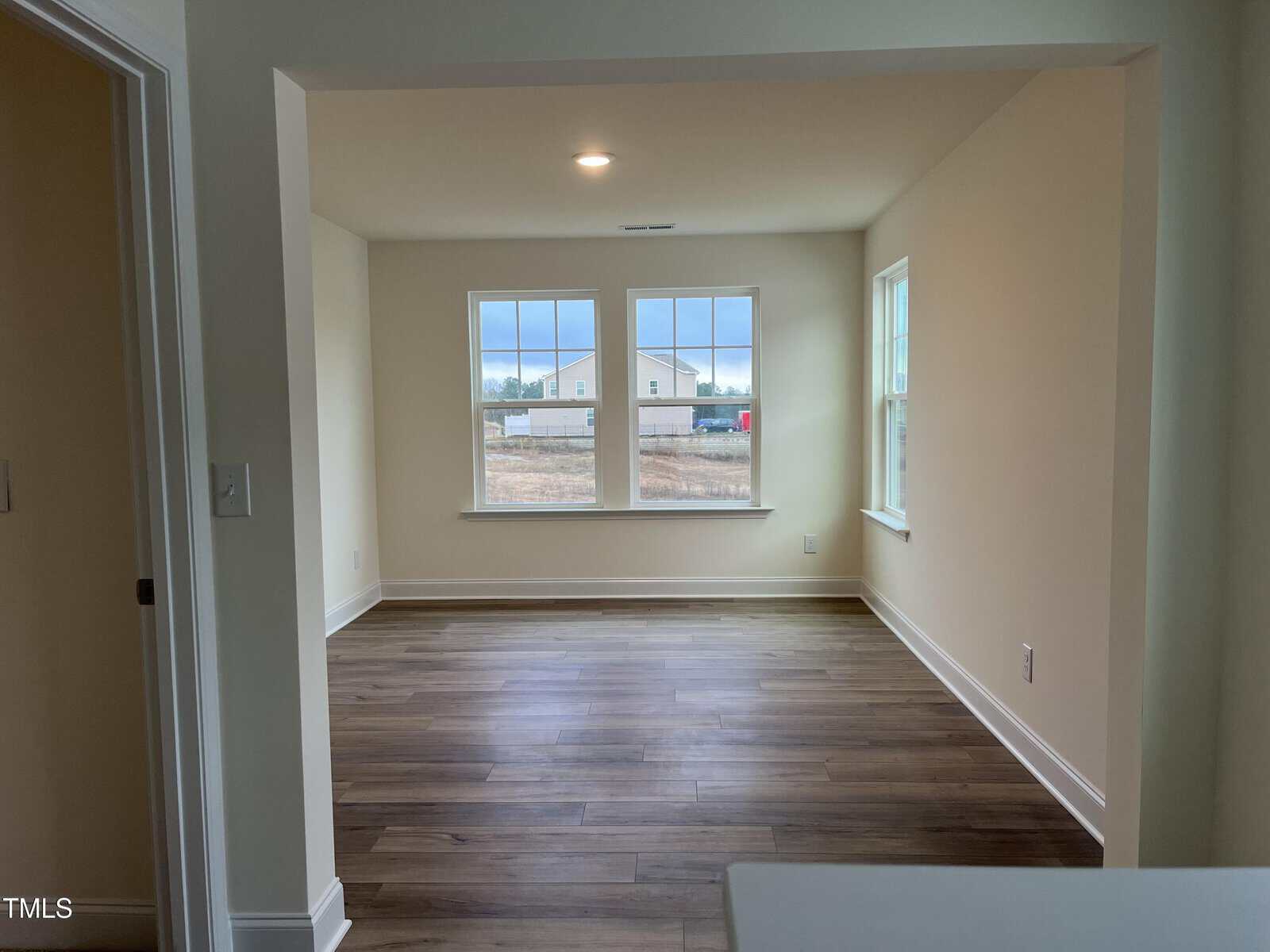Empty room with beige walls, large windows showing an outdoor scene, and dark wood flooring. Sunlight brightens the space, and the room appears clean and unfurnished. Trim is white, and ceiling has a recessed light.