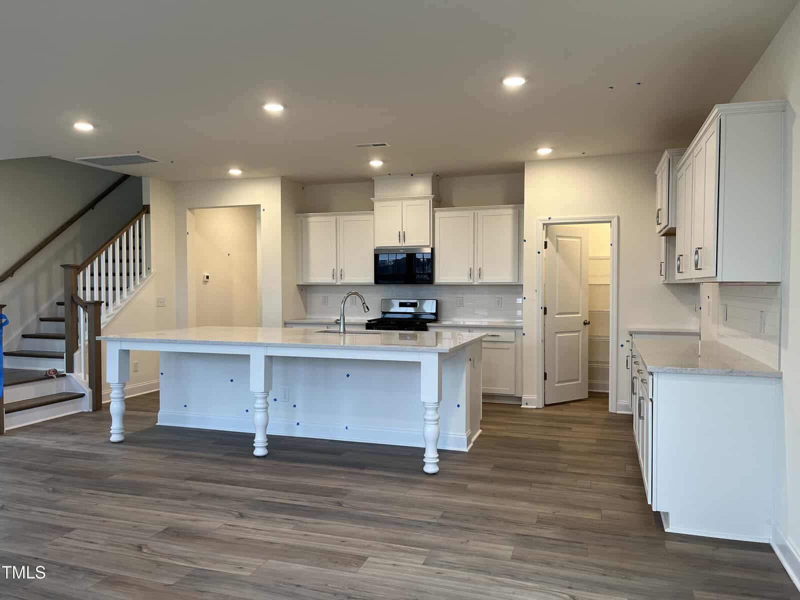 Modern kitchen with white cabinets, stainless steel appliances, a large island with a sink and pendant lights, wood-style flooring, and a staircase visible to the left. Walls are light-colored, giving a bright and open feel.