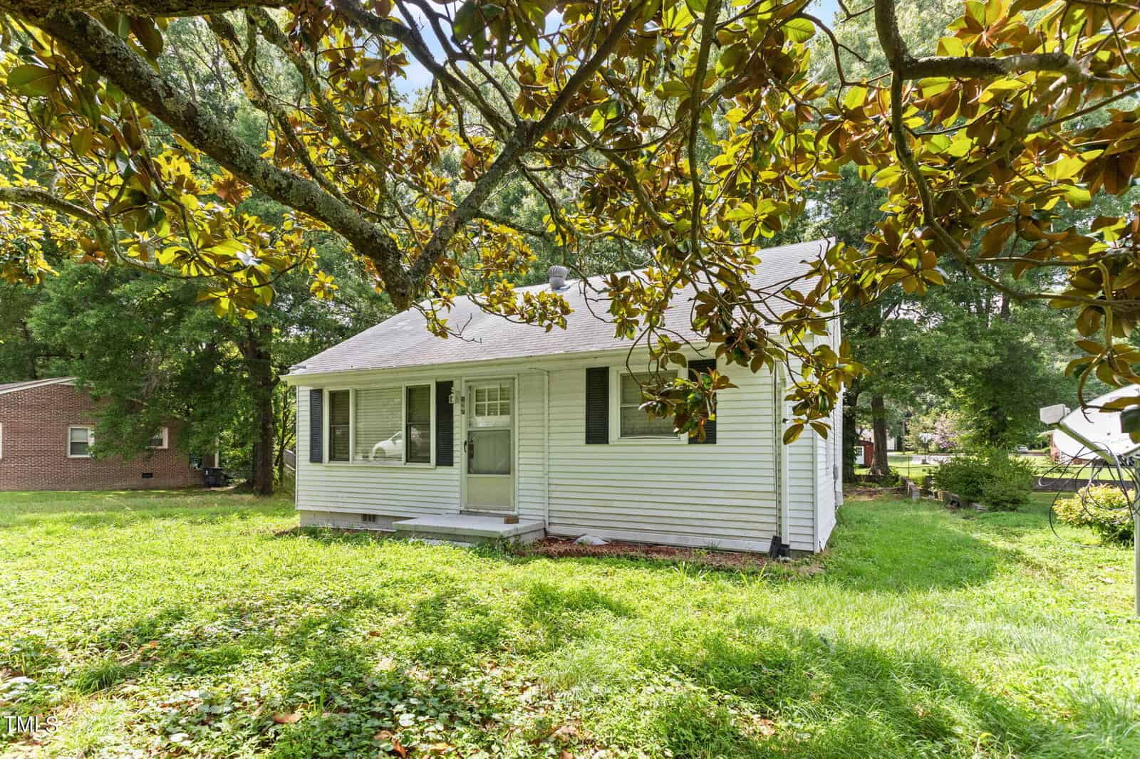 A small, white, single-story house at 127 N Bullock Street in Henderson, with a gray shingled roof, partially obscured by tree branches with green leaves. The house features a front door with a small porch and is surrounded by a grassy yard. A brick building is visible in the background.