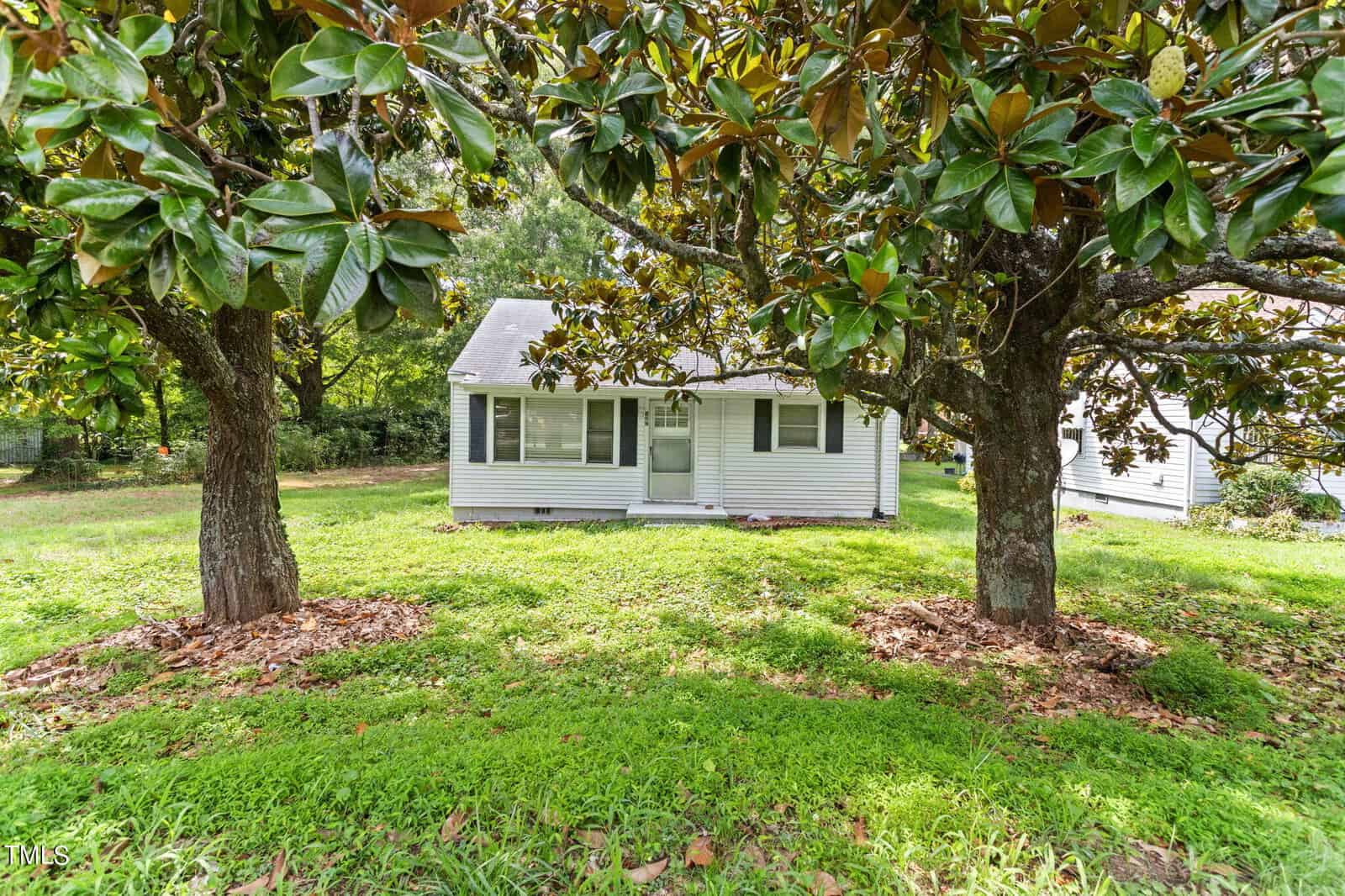 A small, white, single-story house is partially hidden behind two large trees with broad green leaves. The scene features lush, well-maintained green grass and additional trees in the background, creating a tranquil, shaded setting.