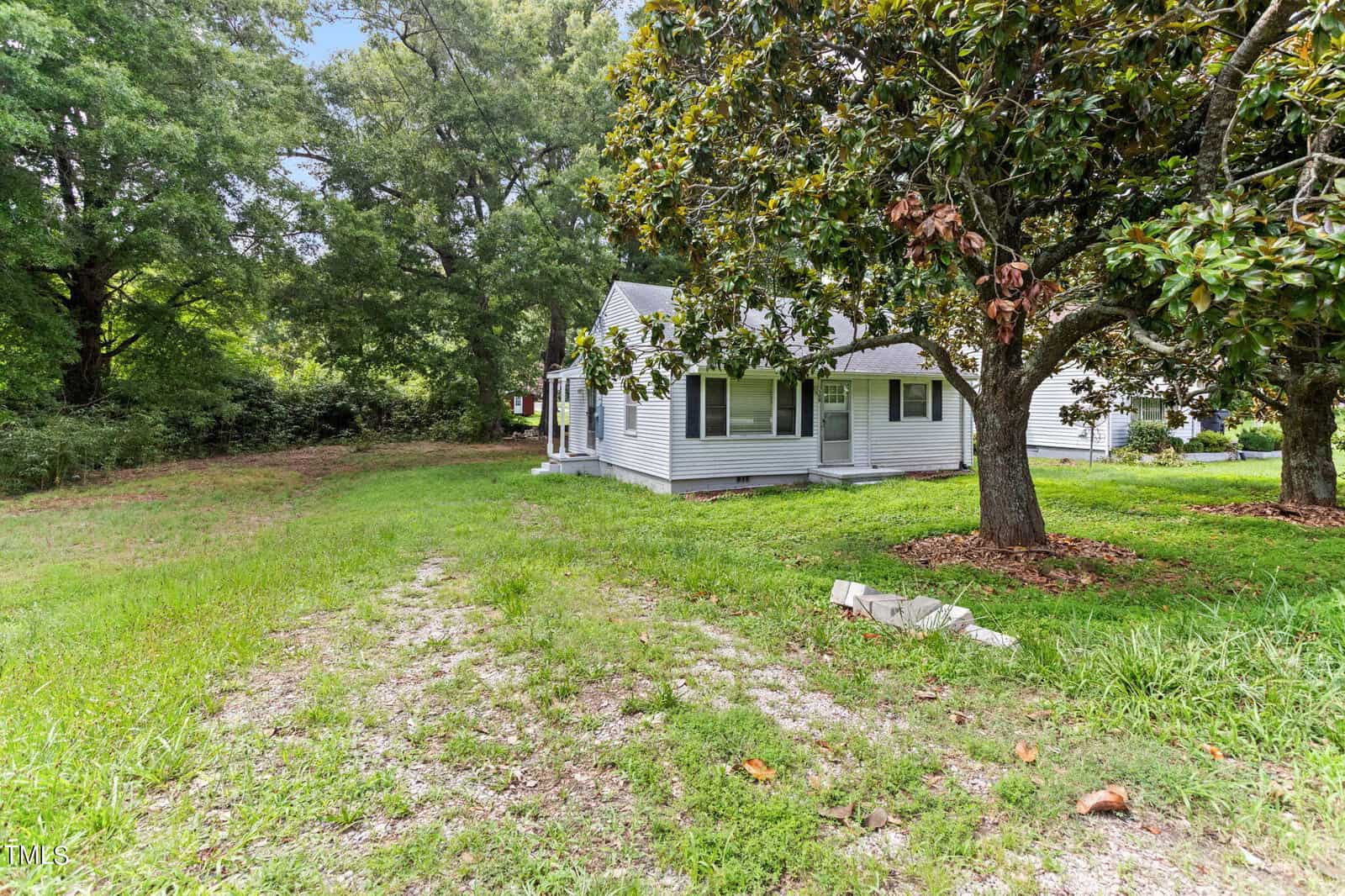 A quaint single-story house with light gray siding is nestled among large, leafy trees. The lush green lawn extends around the property. Natural sunlight filters through the trees, creating a serene and welcoming atmosphere.