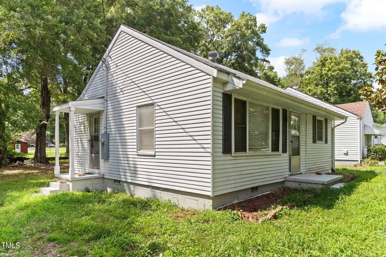 A small, single-story house with light-colored siding and a gable roof is shown. The house features a small covered front porch and several windows along its sides. It is surrounded by a grassy lawn with large trees in the background under a partly cloudy sky.