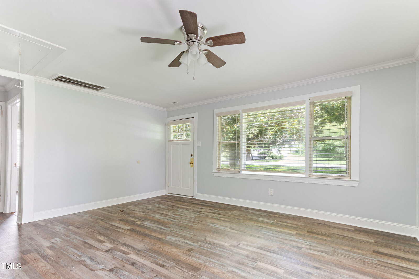 A bright and empty living room with light grey walls and wood flooring. The room features a ceiling fan with lights, a large window with white trim and blinds offering a view of greenery outside, and a white front door with a glass panel.