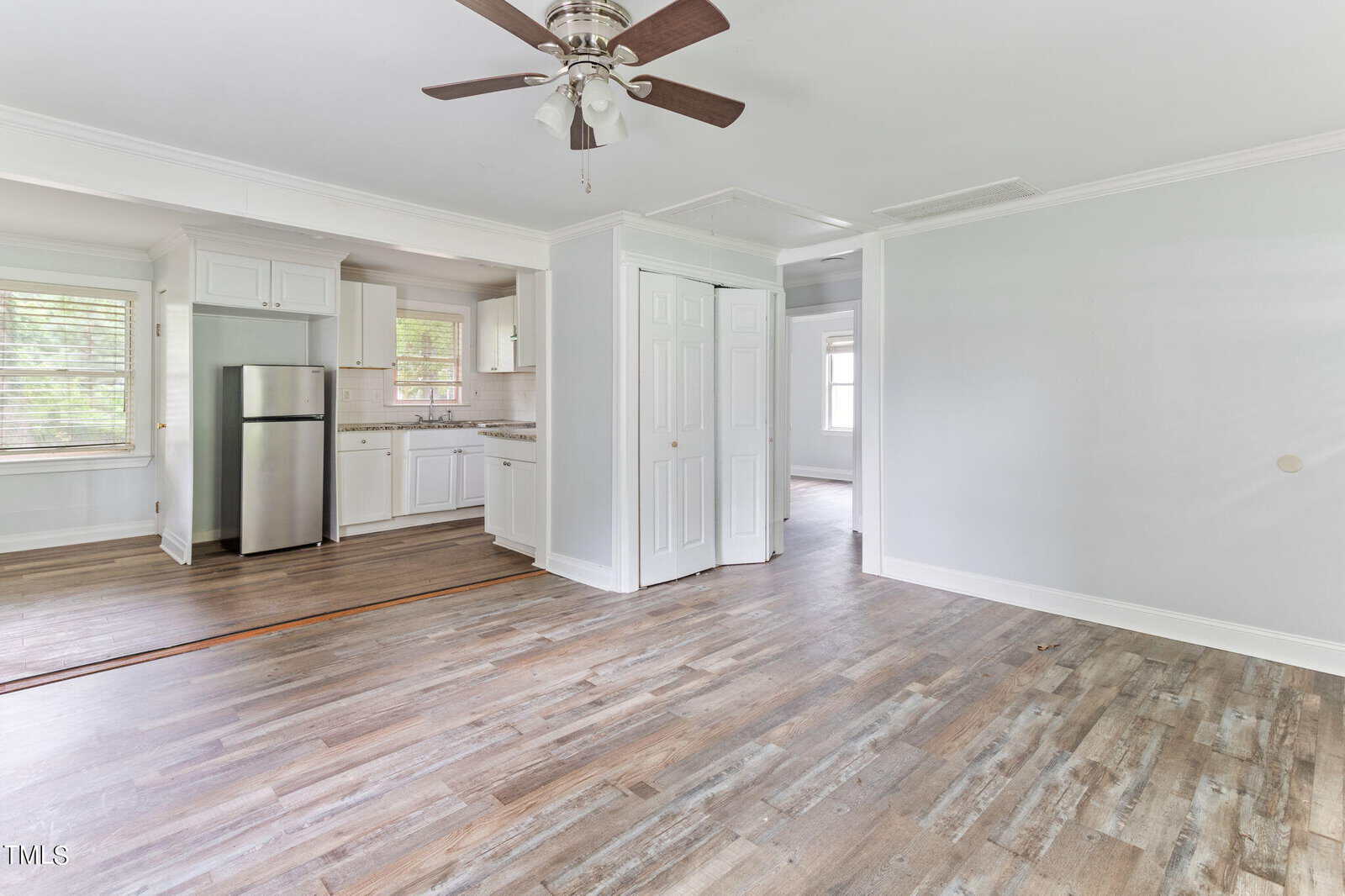 A spacious room with light-colored walls and wooden flooring. It has a ceiling fan, a small kitchen with white cabinets, and stainless steel appliances on the left. A doorway leads to another room in the background, and there are multiple windows letting in natural light.