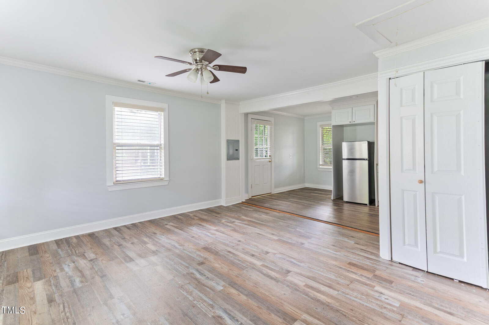 A bright room with light gray walls, white trim, and a ceiling fan. The space has wood-look flooring and several windows. To the right, there is a small kitchen area with a stainless steel refrigerator. A door leads to another room.