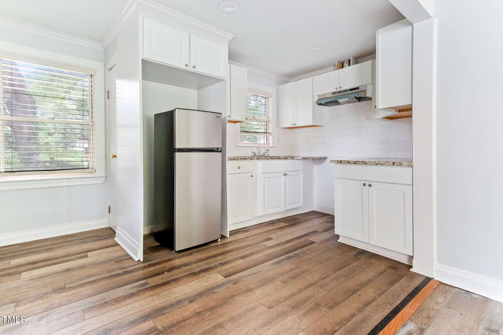 A small, modern kitchen with white cabinets, wood flooring, and a stainless steel refrigerator. A window with blinds allows natural light in, and there is a range hood above the stove area. The countertop has a granite finish and the backsplash is white tile.