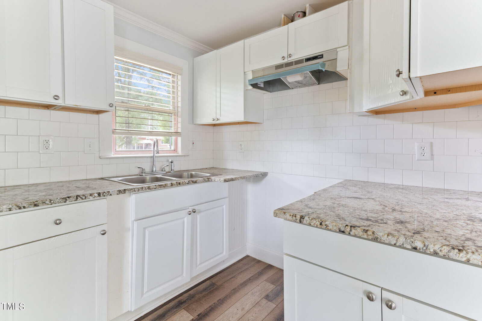A bright kitchen with white cabinets and light gray granite countertops. The room features a stainless steel range hood, a double sink beneath a window with white blinds, and a subway tile backsplash. The floor is covered with light wood plank flooring.
