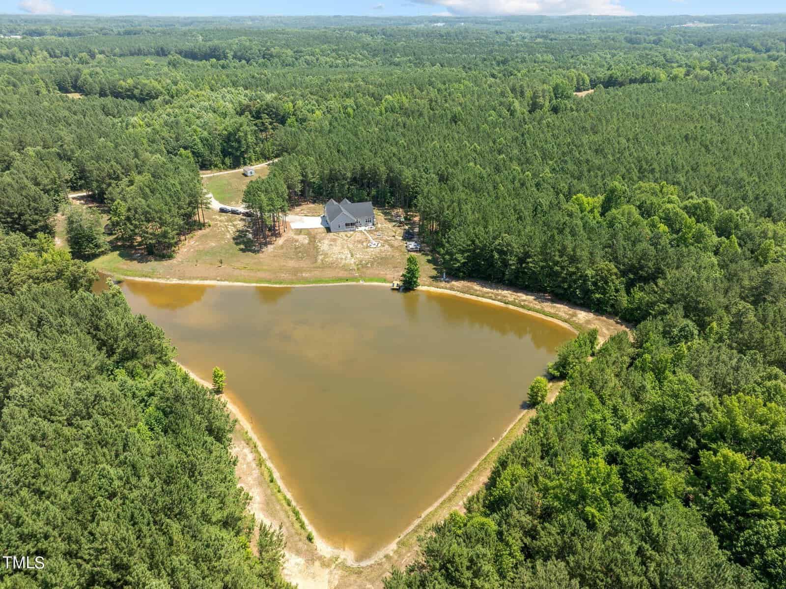 Aerial view of a triangular pond surrounded by dense forest. At the corner of the pond is a large, modern house with multiple outbuildings and a driveway extending through the trees, leading to the property. The landscape is lush and green under a partly cloudy sky.