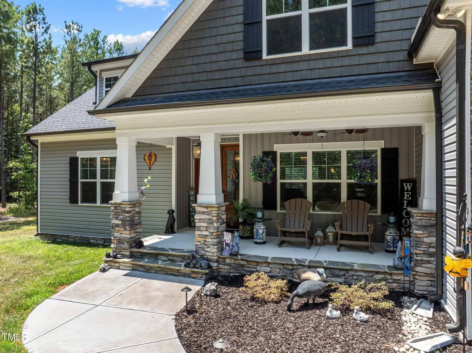 A house with a well-maintained front porch featuring stone pillars, two Adirondack chairs, potted plants, and a "Welcome" sign. The lawn is neatly trimmed, and there are garden beds with mulch and decorative items in the foreground. Trees and blue sky in the background.