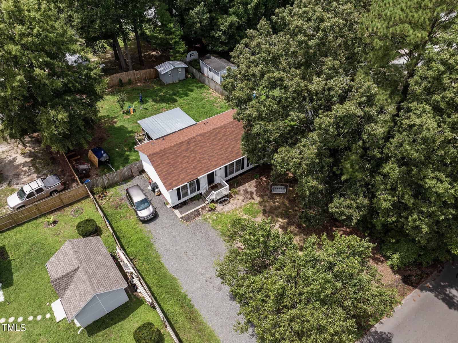Aerial view of a small house with a brown roof. The property is surrounded by trees and has a gravel driveway with a parked car. The backyard contains a shed and is fenced. A well-kept lawn and additional structures are visible nearby.