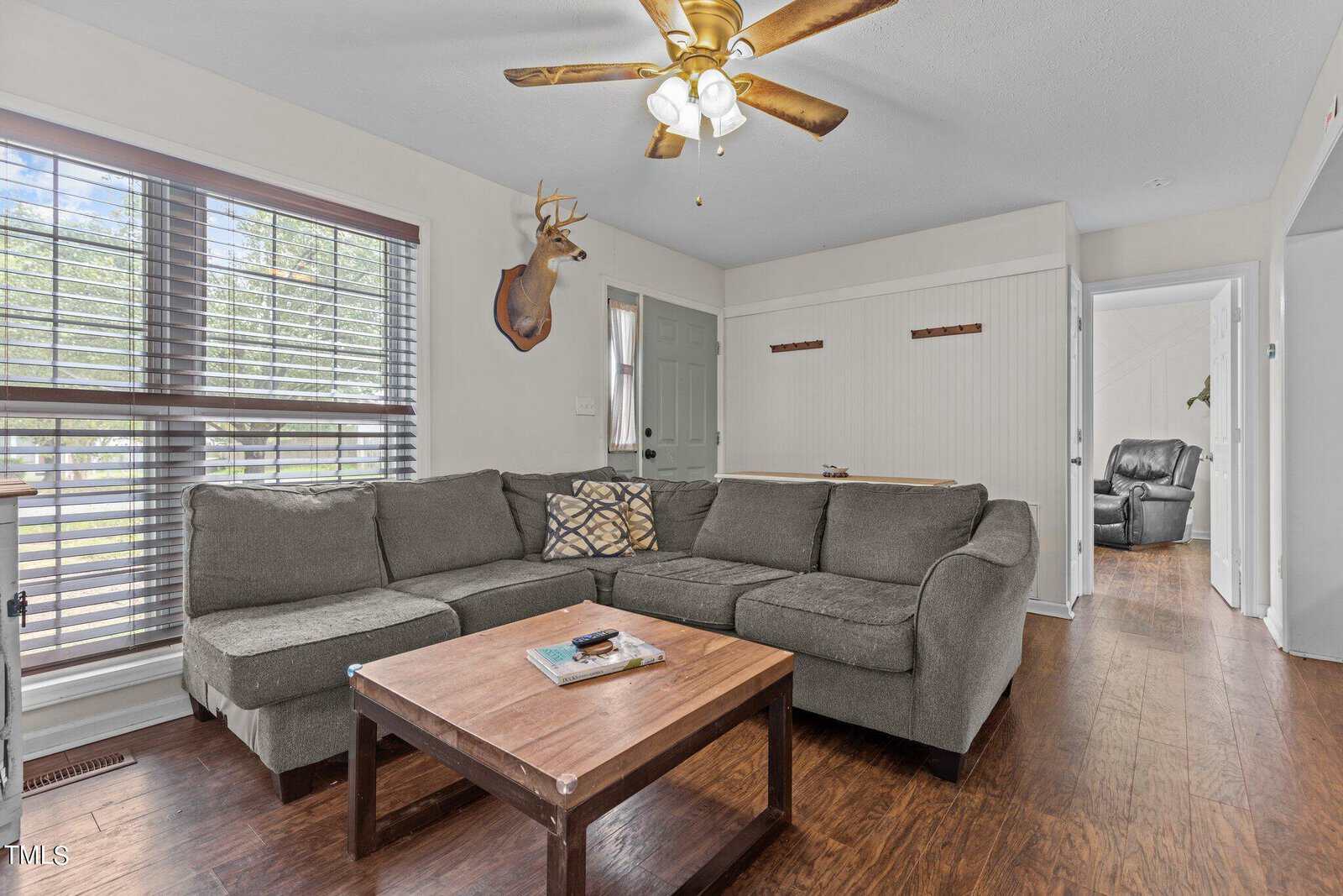 A cozy living room with a large gray sectional sofa and a wooden coffee table. A ceiling fan hangs above, and a mounted deer head decorates the wall. Sunlight filters through the window blinds, creating a warm and inviting atmosphere.