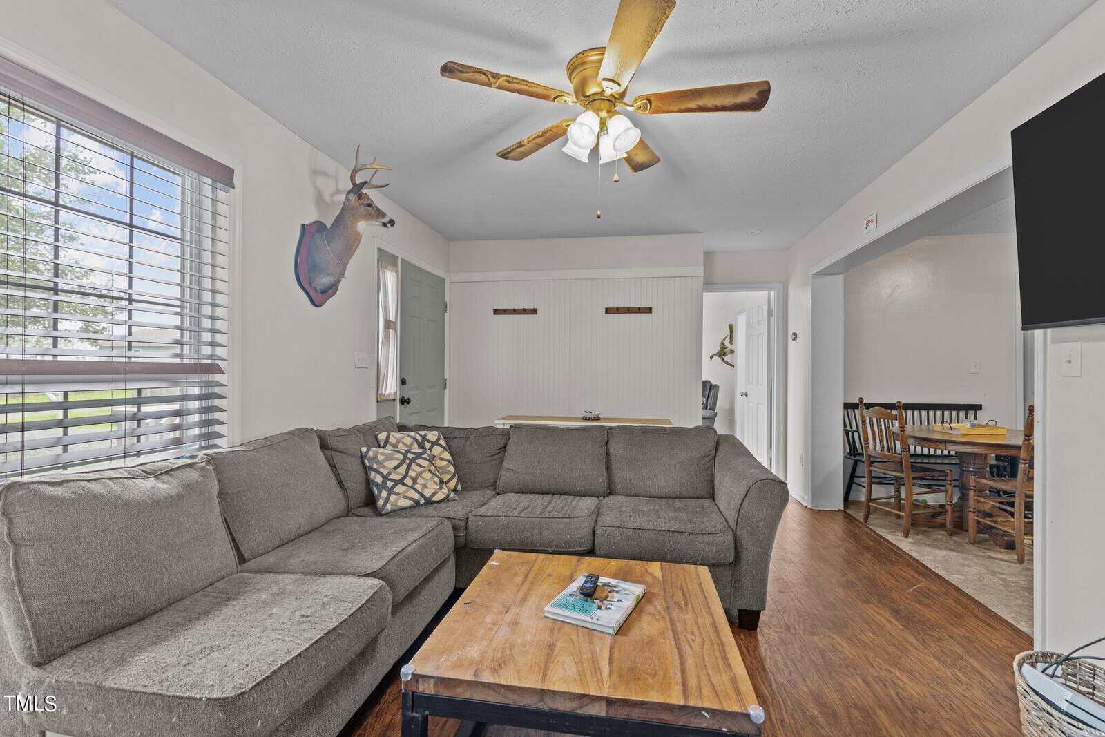 A cozy living room with a gray sectional sofa and a wooden coffee table. A ceiling fan with lights hangs above. A dining area with a table and chairs is visible through an open doorway. A mounted deer head decorates the wall.