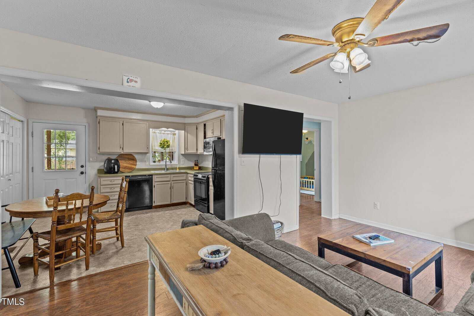 A cozy living space featuring a wooden dining table with four chairs, a modern kitchen with beige cabinets and black appliances, a comfortable seating area with a gray sofa, a wall-mounted TV, and a ceiling fan. The floor is wooden.