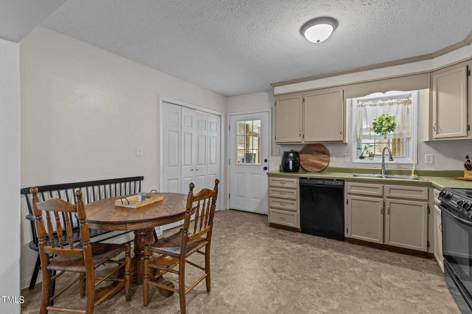 A cozy kitchen with cream-colored cabinets and a light green countertop. A round wooden table with four matching chairs sits nearby. The room includes a stove, oven, dishwasher, and a door with windows. A small vase with flowers decorates the window.
