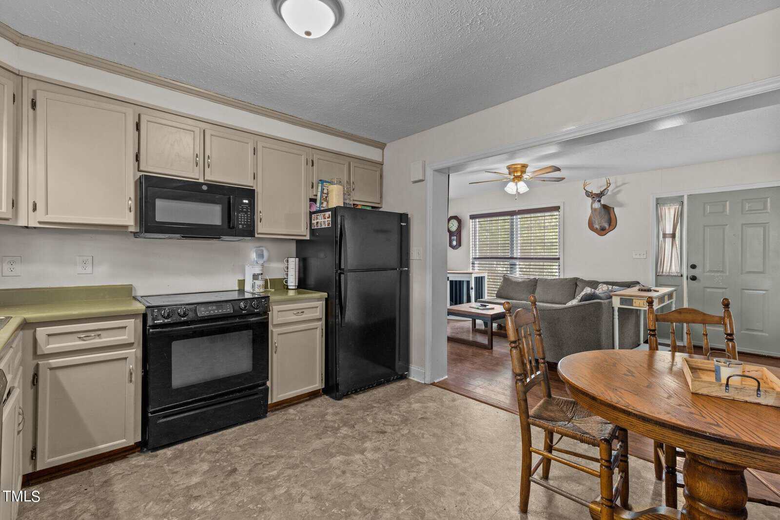 A cozy kitchen-dining area with light cabinets, a black fridge, black stove, and microwave. A wooden dining table with chairs is in the foreground. In the background, a living room with a fan, couch, and mounted deer head is visible.
