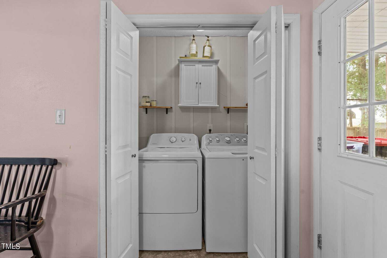Small laundry nook with a washer and dryer inside white double doors. Shelves hold detergents and a small cabinet above the machines. A door opens to the outside, and a black chair is positioned to the left. Natural light filters in from a window.