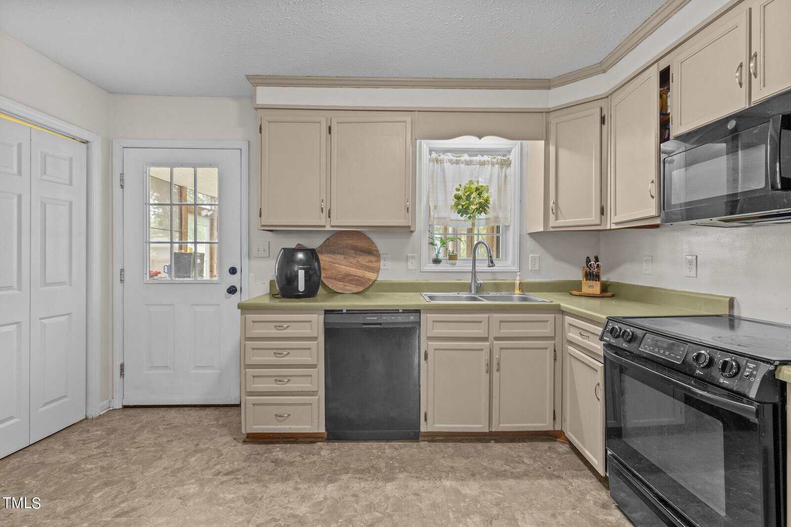 A kitchen with light-colored cabinets, a black dishwasher, and a matching oven. Countertops hold a cutting board, an air fryer, and a knife block. A window above the sink lets in light, and a door with glass panes is on the left.