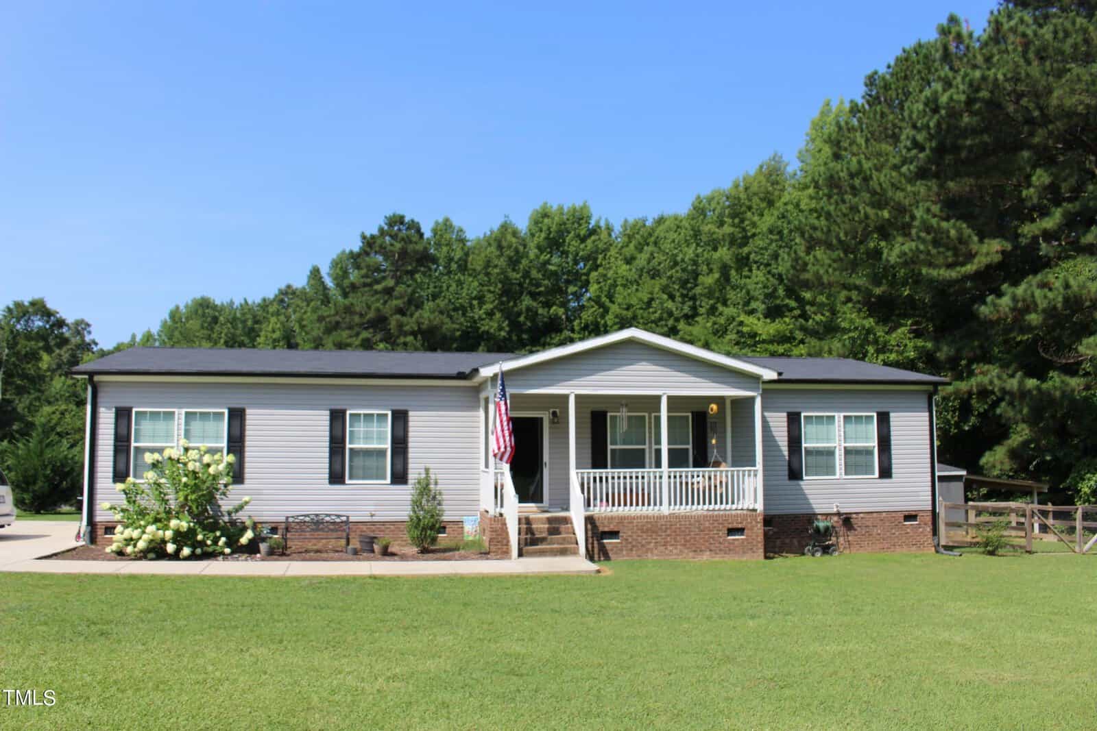 A charming single-story home at 1690 Tally Ho Road in Oxford, with light gray siding and a dark roof, features a front porch with brick steps. An American flag waves by the entrance, surrounded by a well-maintained lawn and lush green trees in the background.