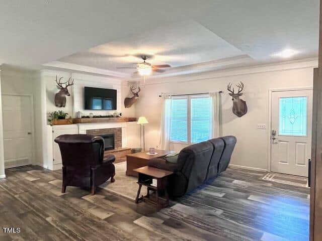 A living room with a ceiling fan, a fireplace, and a mounted television. Two armchairs face a couch, and deer heads are mounted on the walls. A door with a decorative window is to the right, and natural light filters through the windows.