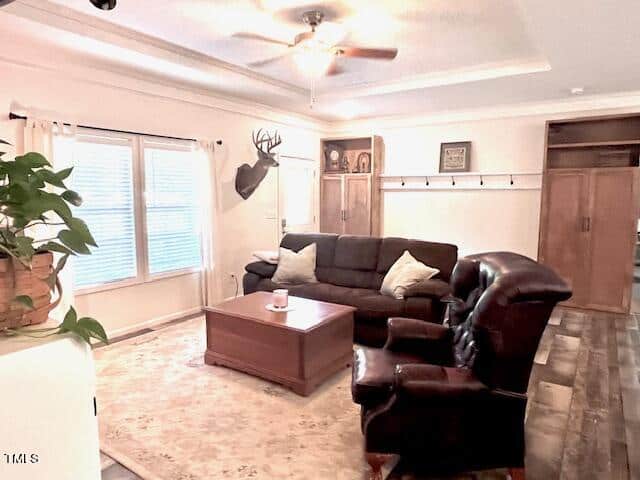 Cozy living room with a dark sofa and two leather chairs, centered around a wooden coffee table on a light rug. A ceiling fan is above, and a deer head mount decorates the wall. Large windows let in natural light, and a potted plant is nearby.