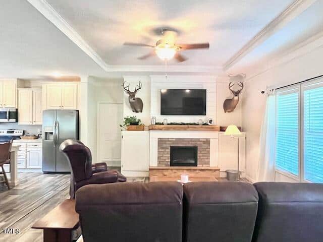 A cozy living room with a brown sofa facing a fireplace topped by a TV. Deer head mounts are on either side of the mantel. A ceiling fan hangs above, and the kitchen with wooden cabinets is visible in the background.
