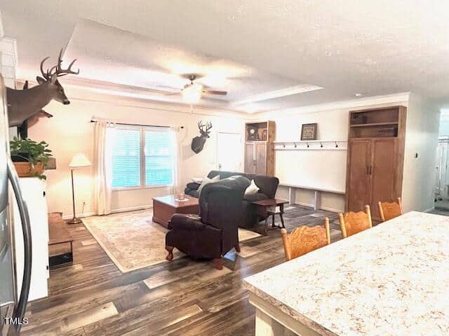 A cozy living room with wood flooring and rustic decor. Mounted deer heads adorn the walls. The room features a large window with closed curtains, a plush armchair, a sofa, and a hardwood coffee table. A ceiling fan is centered above the seating area.