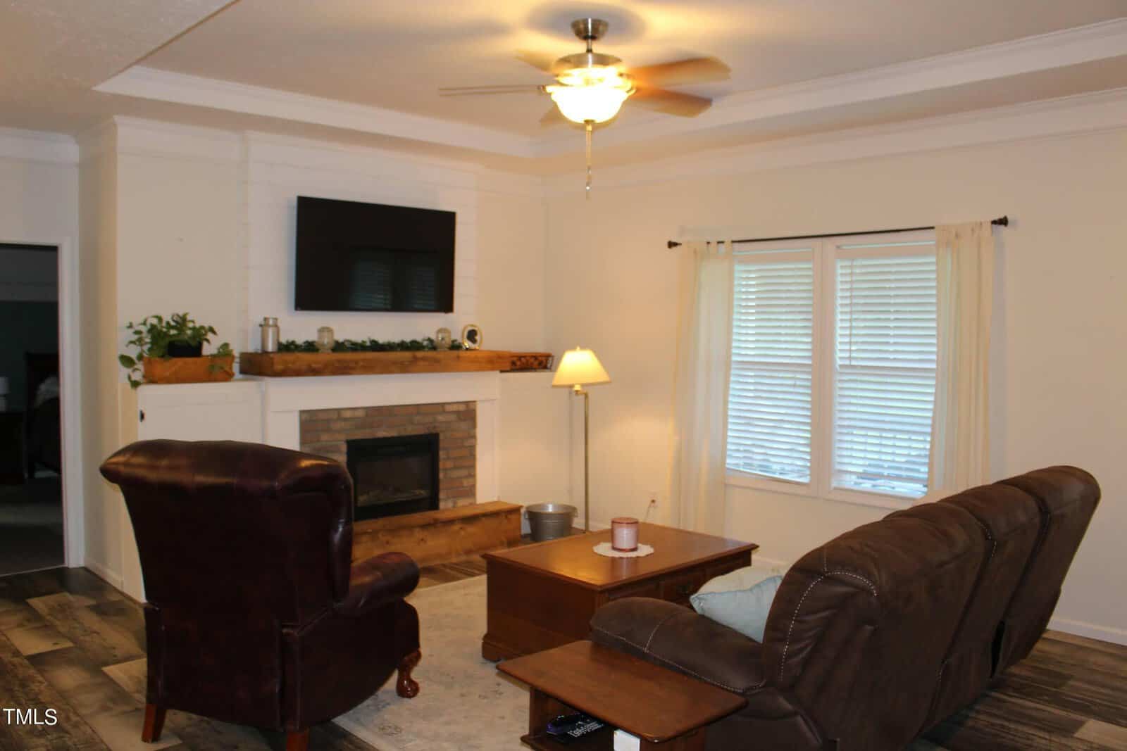 A cozy living room with a brown leather armchair and a sofa facing a brick fireplace. A flat-screen TV is mounted above the mantel, which is decorated with plants. A lamp and a window with curtains add warmth to the space.