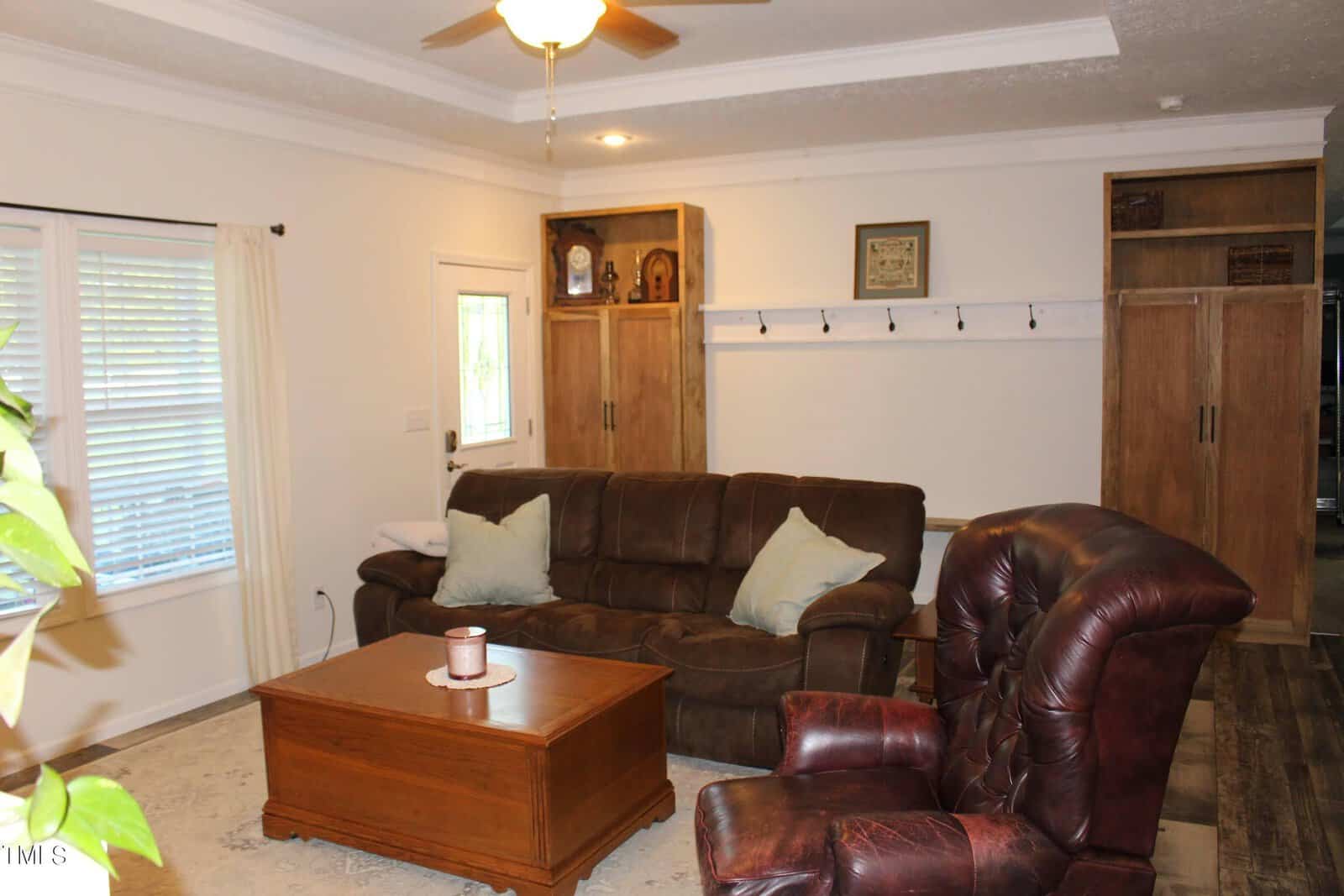 A cozy living room with a brown leather sofa and recliner, a wooden coffee table, and built-in wooden cabinets. A ceiling fan hangs overhead, and two windows with white curtains provide natural light. A small candle sits on the table.