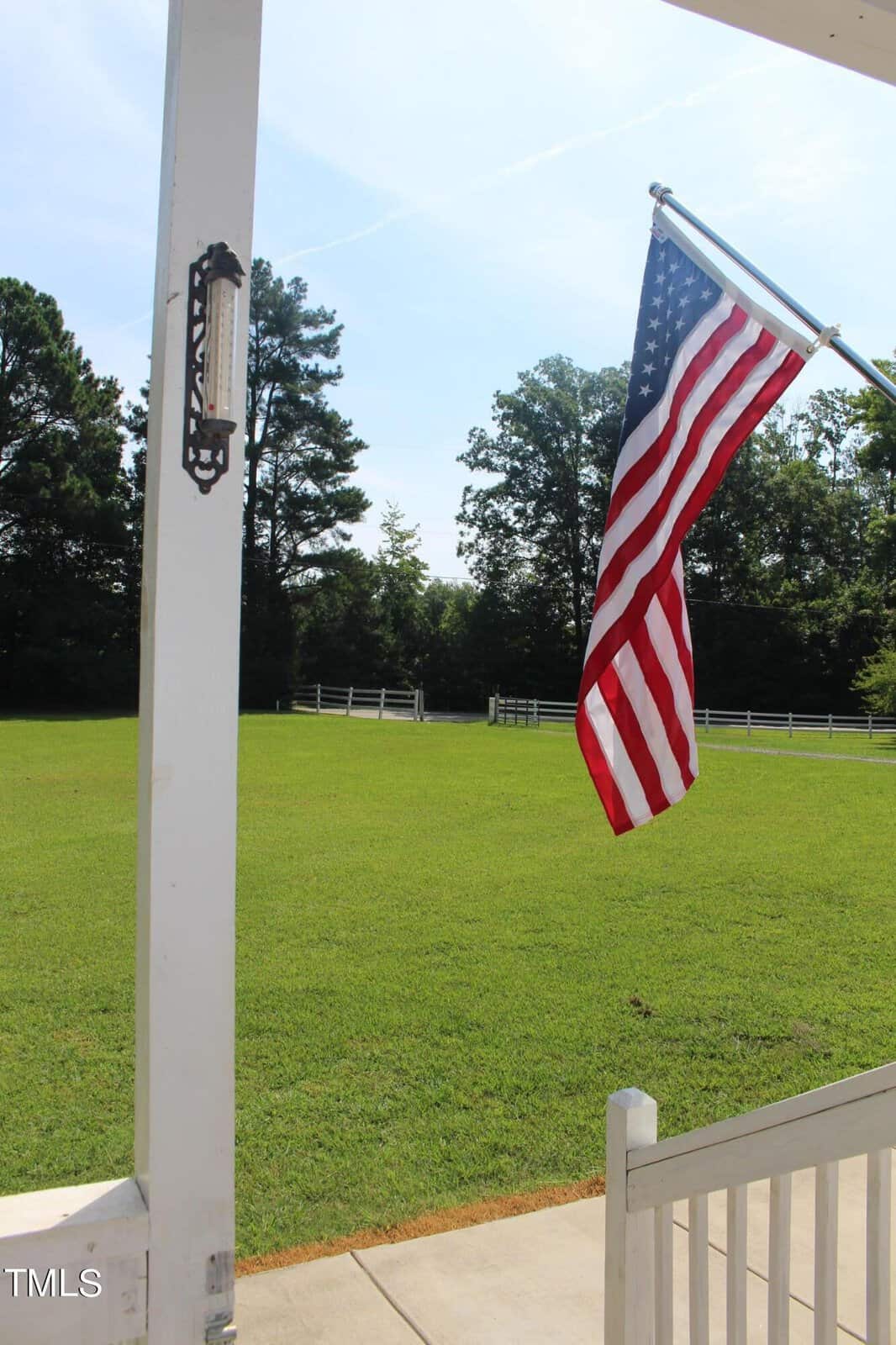 American flag flying on a porch overlooking a green lawn with trees and a white fence in the background.