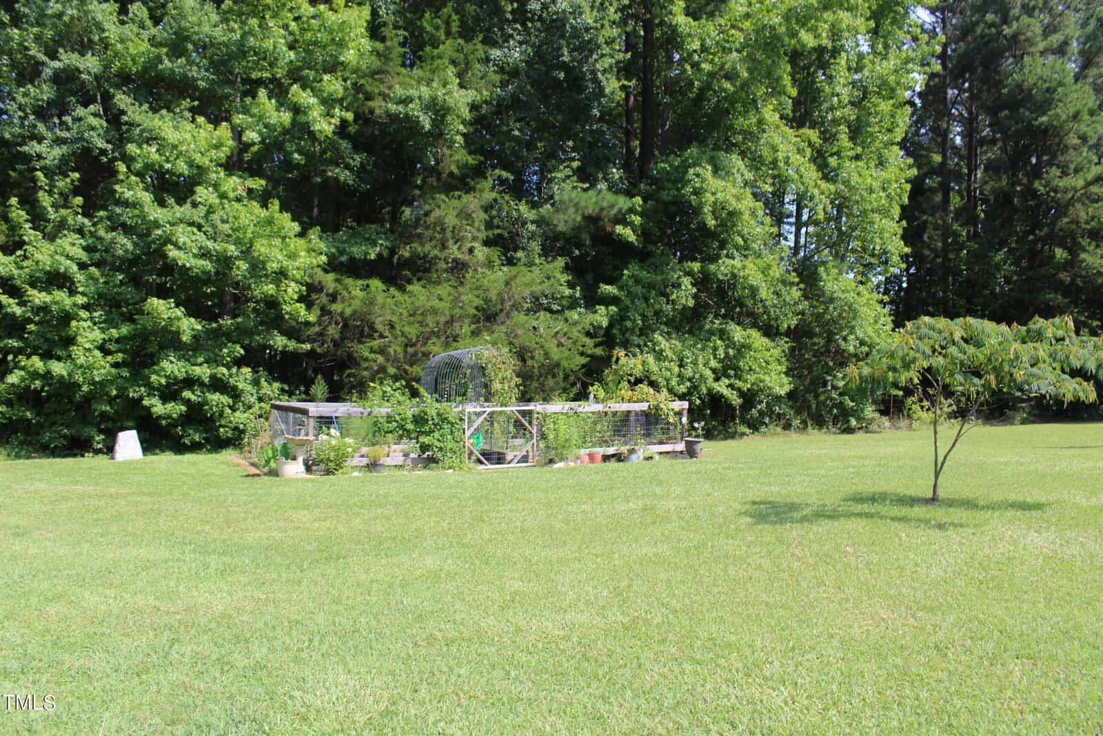 A small garden enclosed with wire fencing is situated in the middle of a grassy field. There are various plants inside the enclosure. Surrounding the garden are dense, green trees under a clear sky.