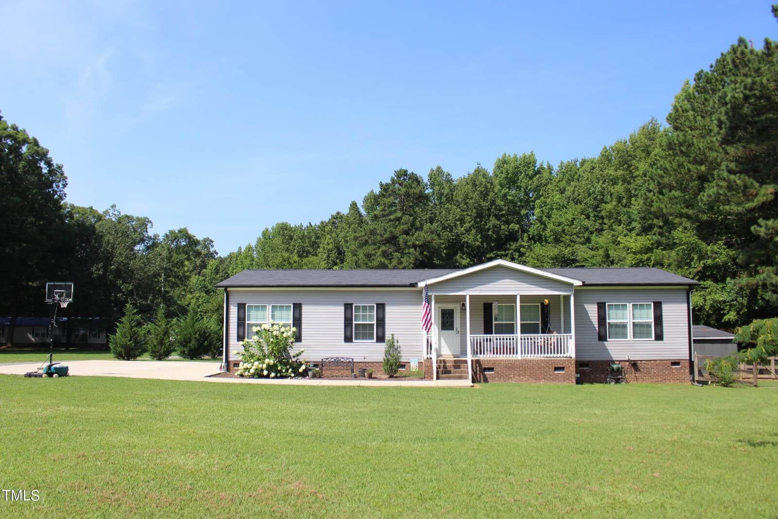 A single-story house with a gray exterior and a small porch, set in a lush green yard. An American flag is displayed on the porch. A basketball hoop is visible on a concrete driveway, and trees surround the property.