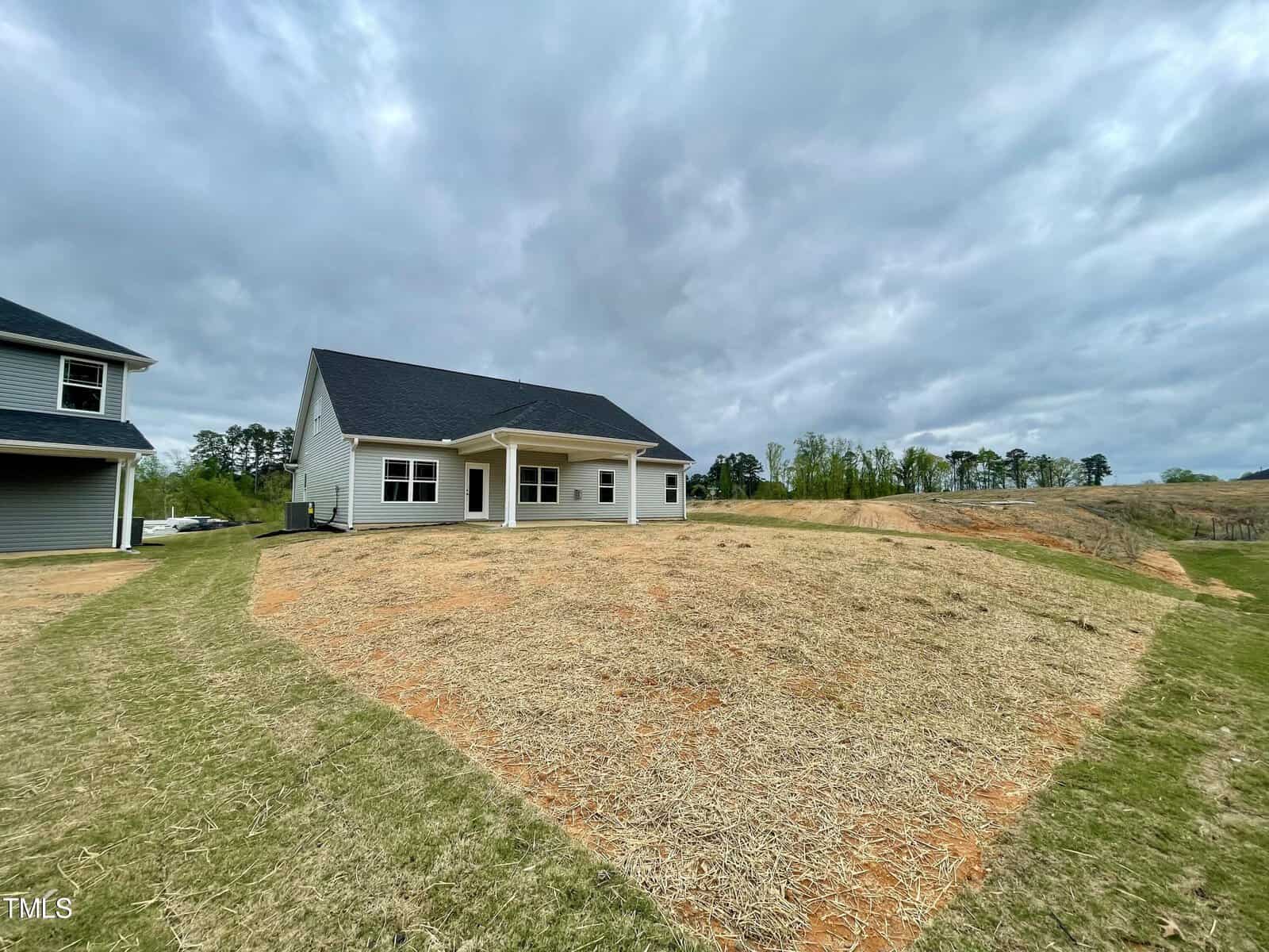 A single-story house with a covered back porch sits on a large, sloped yard with straw-covered ground and patches of grass under a cloudy sky. Trees and open land are visible in the background.