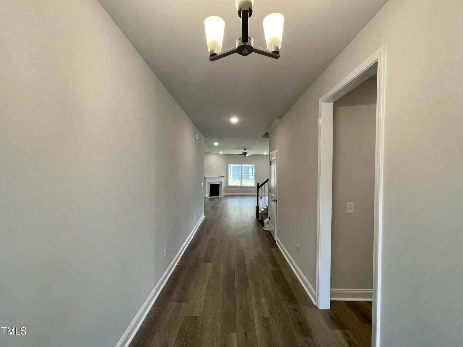 A long hallway with light gray walls, wood flooring, and a ceiling light fixture leads to a living area with a fireplace, ceiling fan, and large window at the far end. A doorway is on the right side of the hall.