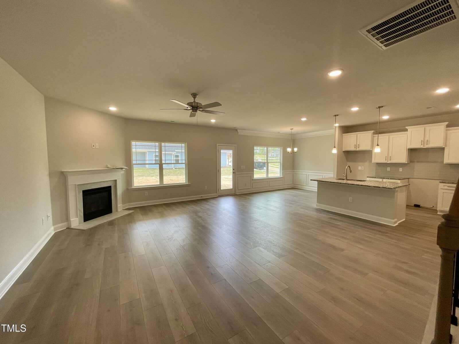 Open-concept living area with light wood floors, a ceiling fan, a corner fireplace, large windows, and a modern kitchen with white cabinets, pendant lights, and a center island.