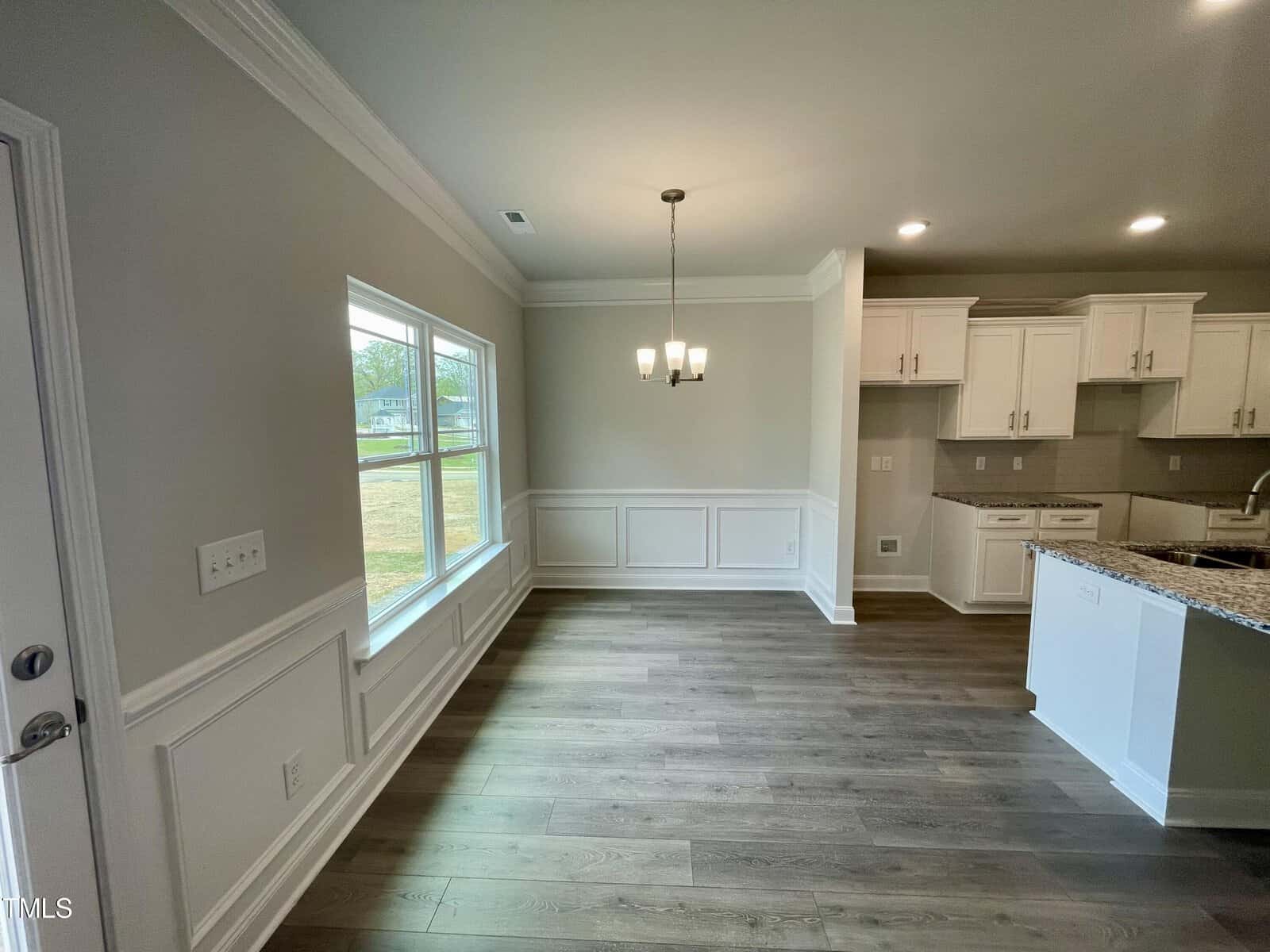 A bright dining area with large windows, light gray walls, wood flooring, a modern chandelier, and an adjacent white kitchen with cabinets and granite countertops.