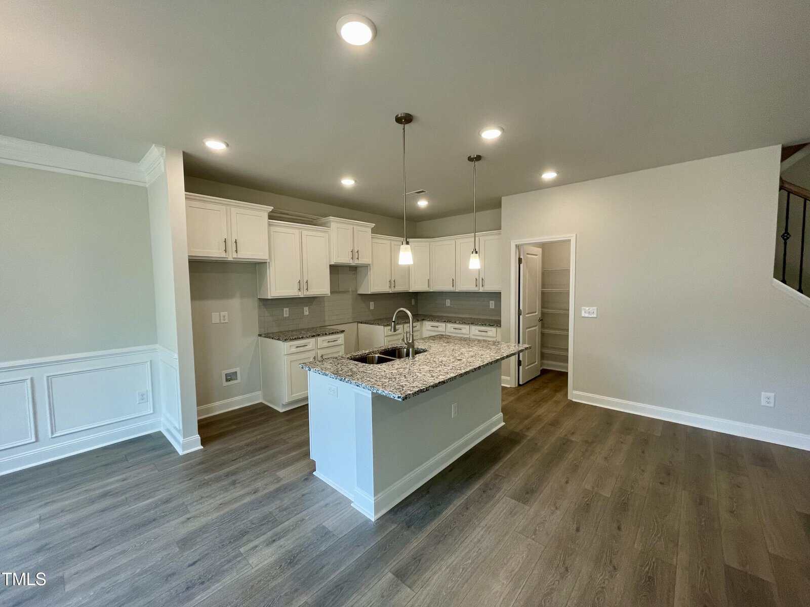 Modern kitchen with white cabinets, granite island with sink, pendant lights, wood-style flooring, and neutral walls. The kitchen opens to a spacious area with wainscoting and recessed lighting. Doorway leads to a separate room.