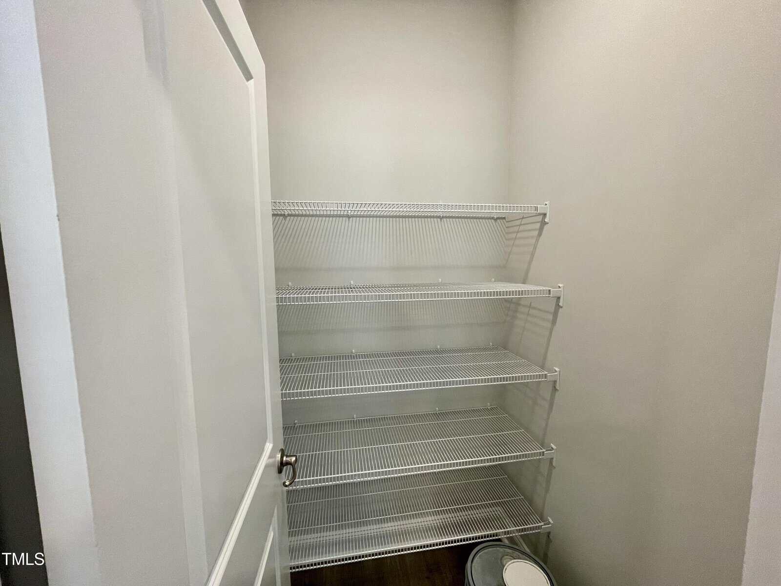 A small, empty closet with four white wire shelves and light-colored walls. The door is open, and a round container is on the dark floor in the corner.