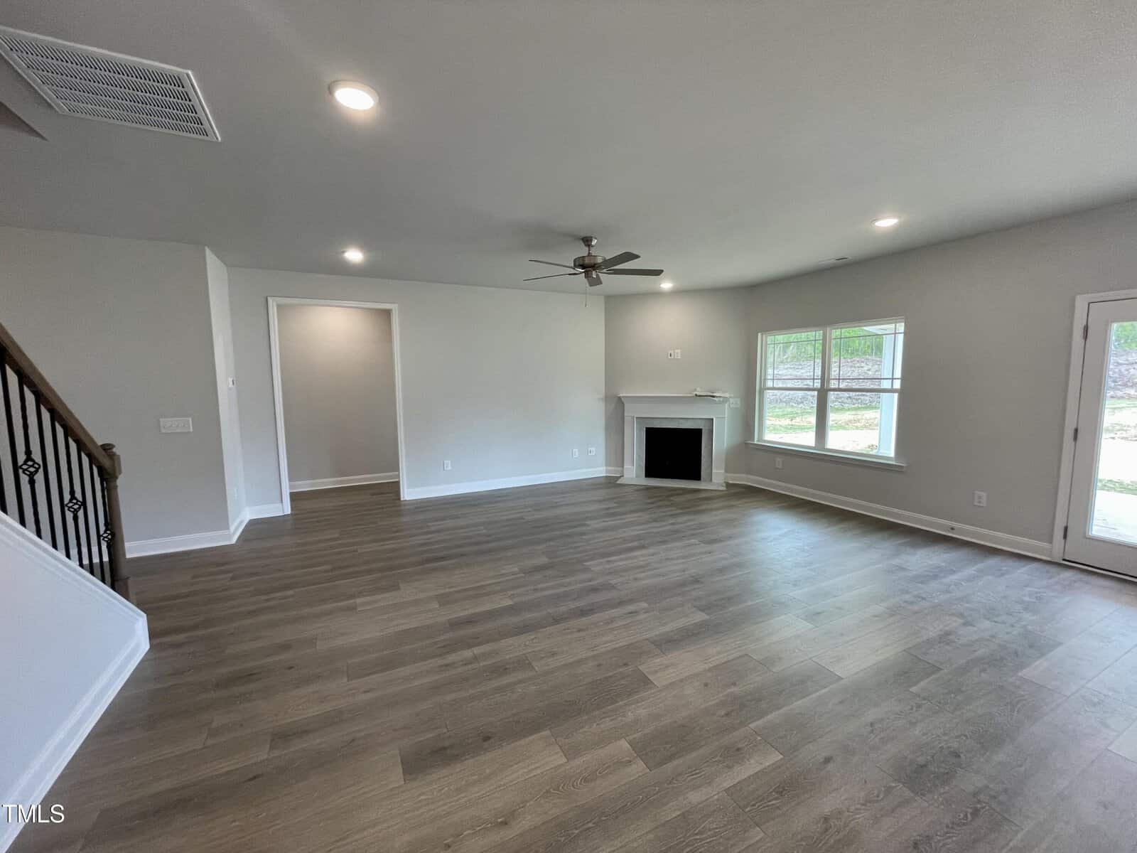 Spacious living room with wood flooring, ceiling fan, recessed lighting, a fireplace, large window, and a glass door leading outside. Stair railing is visible on the left. Walls are painted light gray.